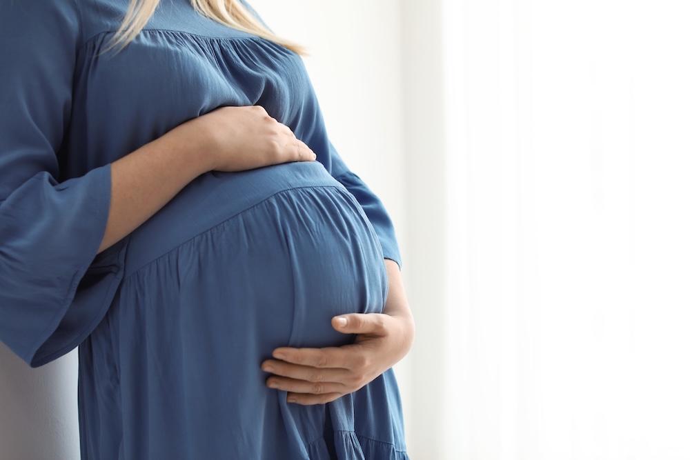 Närbild av en gravid kvinna i blå klänning med händerna på magen.