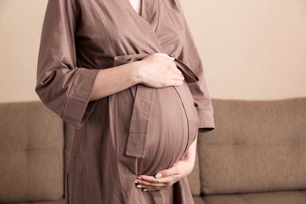 Gravid kvinna i brun klänning håller en hand ovanför och en hand under magen.
